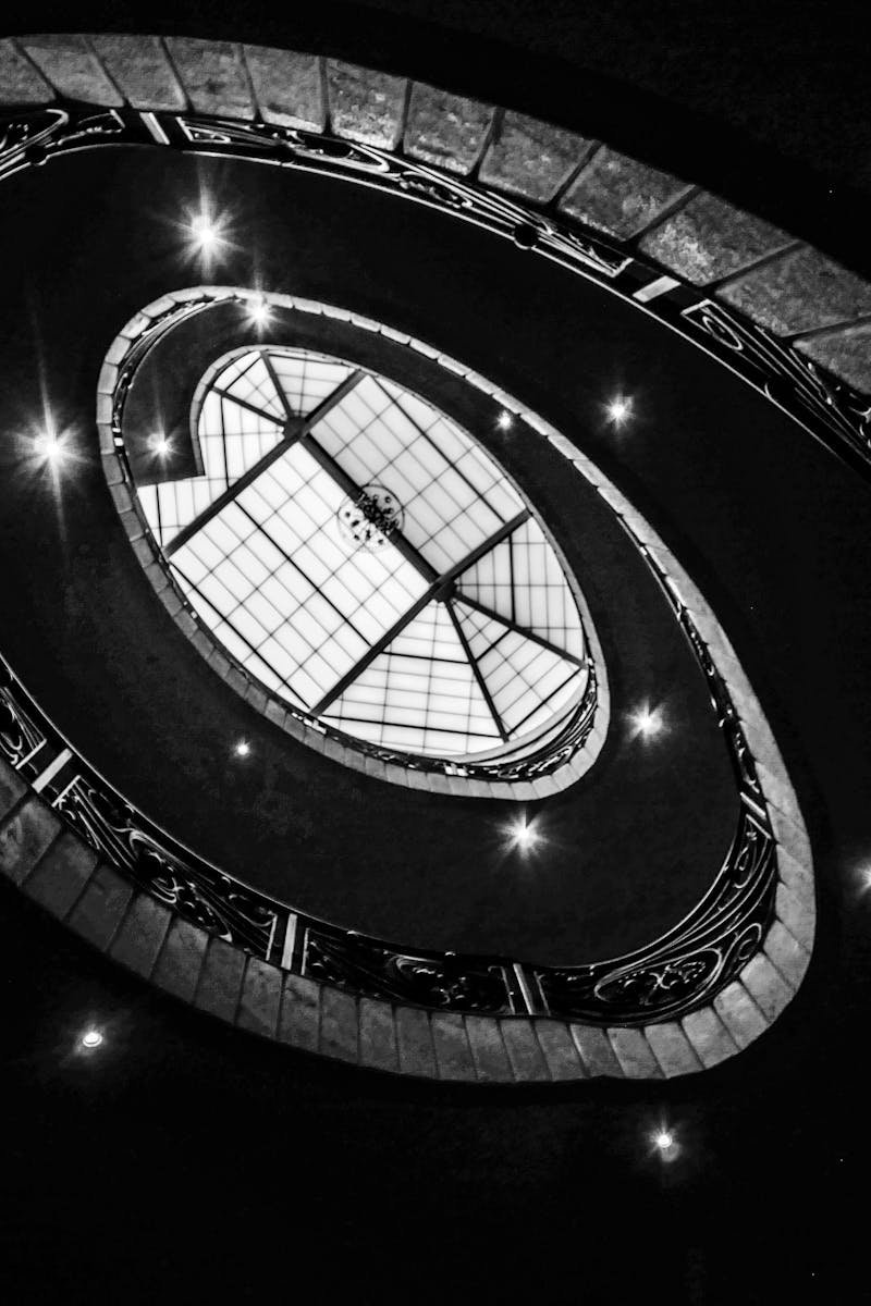 Abstract view of a spiral staircase leading to a geometric skylight, captured in black and white.